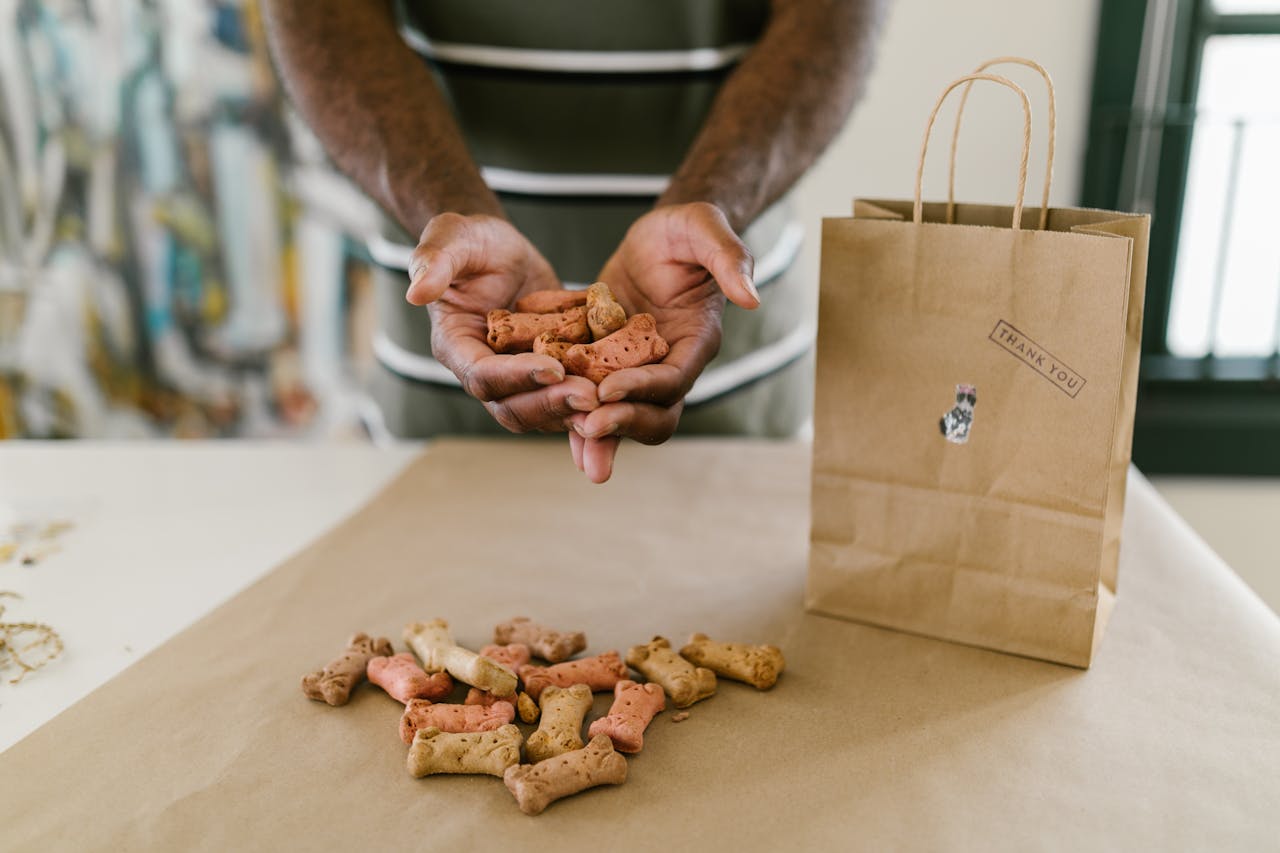 Hands holding colorful dog treats next to a thank you paper bag on a table.